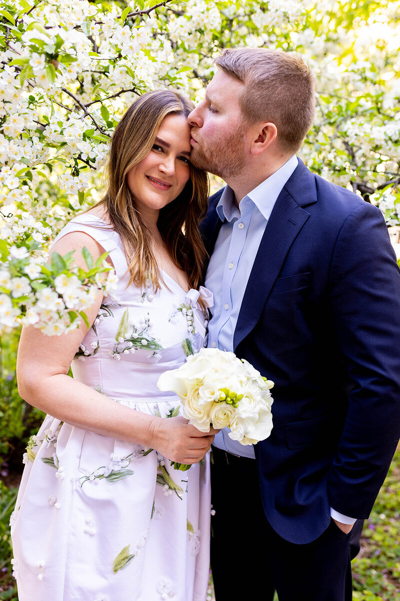 groom kissing the brides forehead outside with  white cherry blossoms behind them