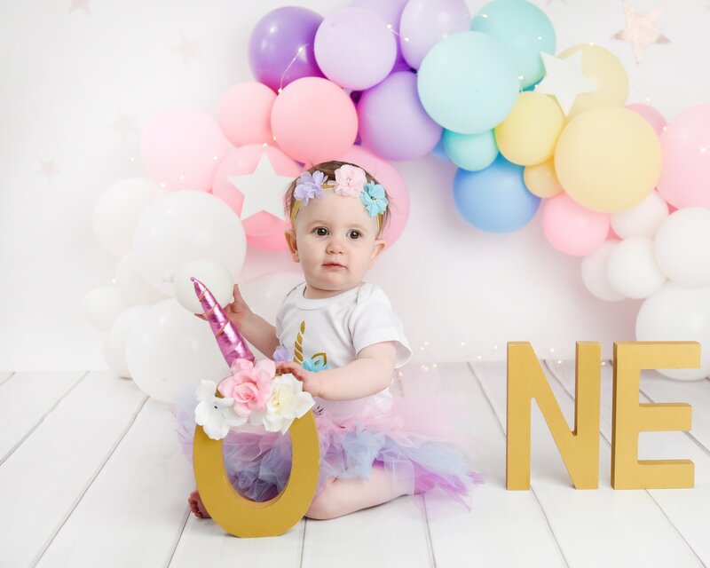 A baby girl wearing a unicorn-themed outfit and floral headband, holding a unicorn "O" decoration with pastel balloon decor in the background.