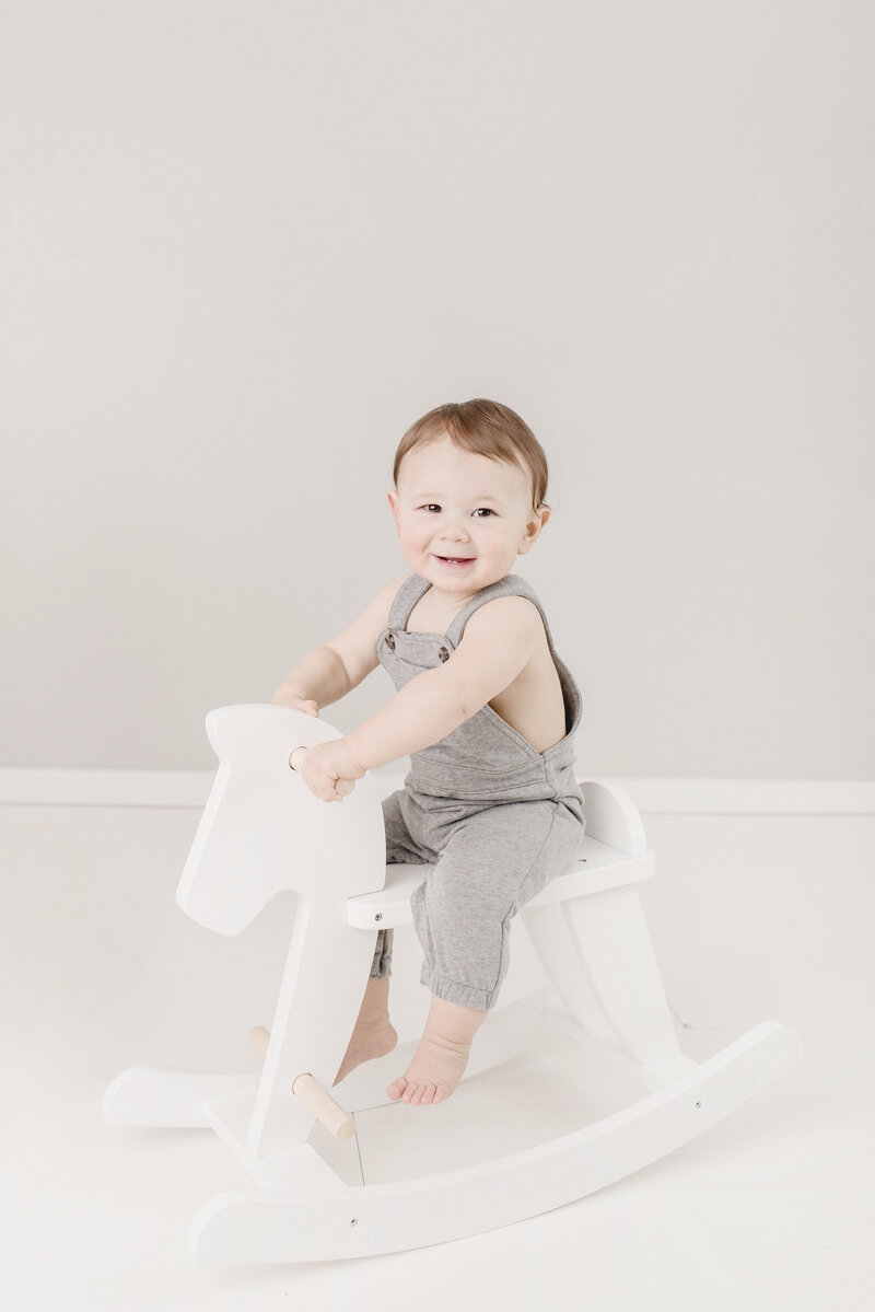 Baby boy in gray overalls riding white wooden rock horse
