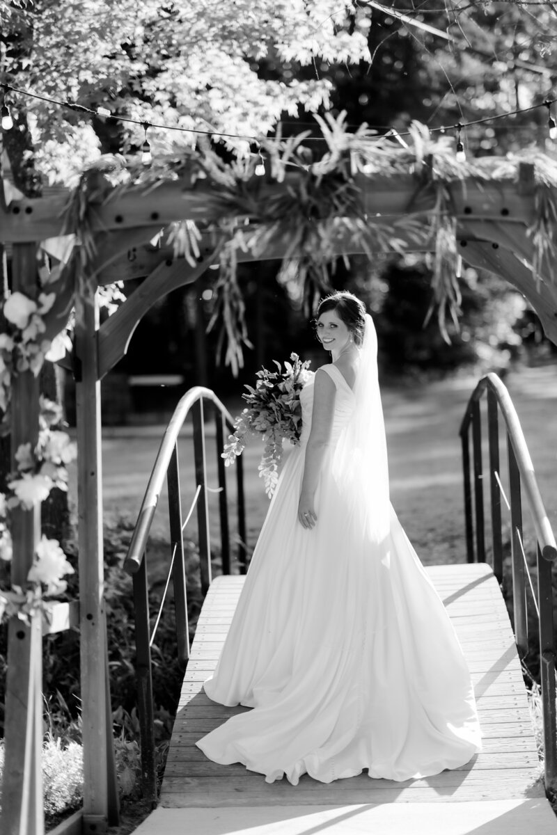 bride walking over a bridge at her outdoor little rock ar wedding venue as she holds her bridal bouquet and looks back at her arkansas wedding photographers