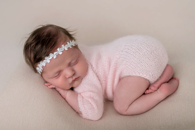 Peaceful sleeping newborn baby girl in pink wrap with white floral headband photographed in classic side-lying pose on cream backdrop