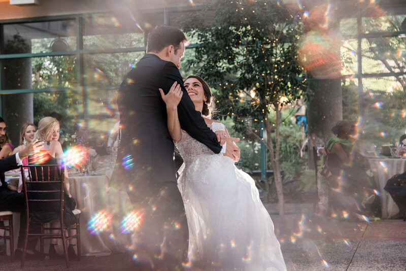white groom in black tux on a dance floor dipping white brunette bride in strapless white gown who is laughing on a dance floor with the light speckling the camera 