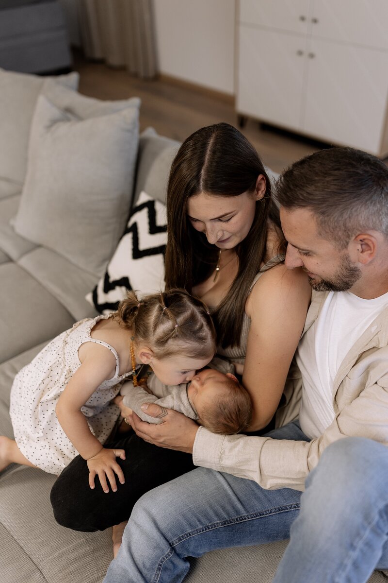 Mutter und Vater sitzen mit Tochter und Baby auf dem Sofa, Tochter gibt Baby einen Kuss