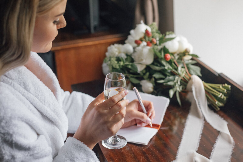 Excited bride shares a glass of champagne and writes down her thoughts ahead of her wedding ceremony