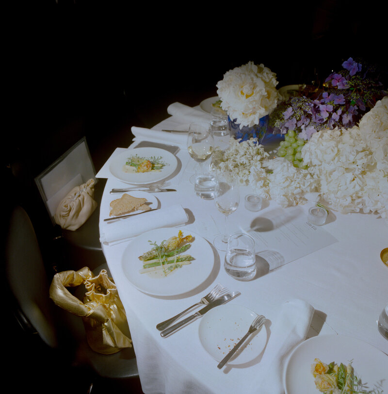 An elegant and minimalistic wedding table setting at Cutler and Co in Fitzroy, Melbourne, showcasing a refined dining experience. The round table is covered with a crisp white tablecloth and features white porcelain plates, each artistically plated with gourmet dishes. The dishes consist of asparagus spears, garnished delicately with edible flowers and herbs, along with thin slices of artisanal bread. Surrounding the plates are neatly arranged wine glasses, water glasses, and polished silver cutlery, adding to the sophisticated ambiance. The table's centerpiece is an opulent arrangement of lush white hydrangeas and purple flowers, beautifully contrasted by clusters of green grapes. A beige purse with a bow detail is draped over one of the black chairs, adding a touch of personality and warmth to the otherwise formal setting. The soft lighting casts subtle shadows, contributing to the overall intimate and luxurious atmosphere of this wedding reception at a high-end venue.