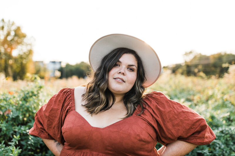 Owner Pam Perez  in rust colored dress and hat back lit by the sun in front of a green and golden field