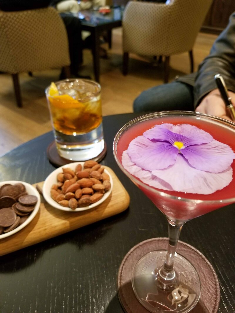 Cocktail garnished with an edible flower, served alongside a whiskey and gourmet snacks in a lounge at The Balmoral, Edinburgh, Scotland ©Stephanie Dosch | theViatrix Luxury Hotel Booking