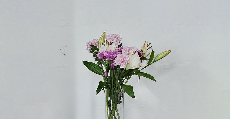 A bouquet of light pink, purple, and white flowers are arranged in a clear vase. The bouquet contains lilies and roses.
