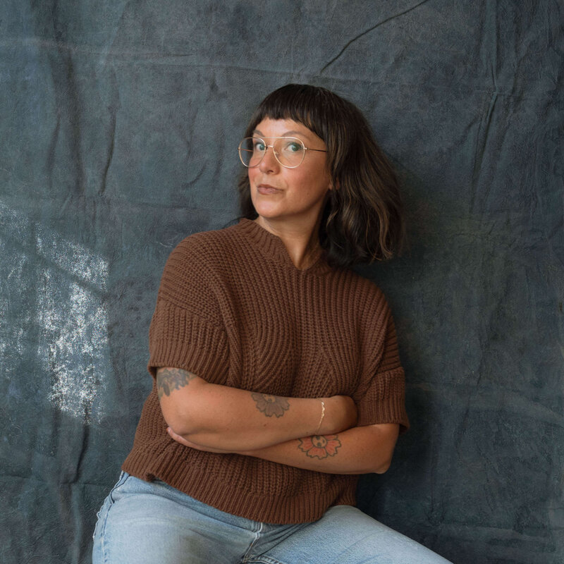 Maria Alcantara, Orange County family photographer, seated against a textured blue backdrop wearing a brown knit sweater and glasses.