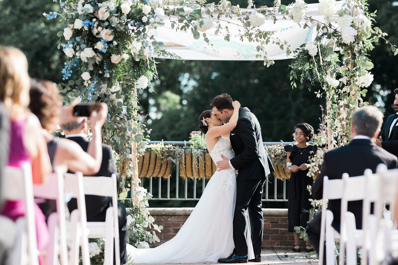 a white groom in a black tux kissing a white jewish brunette bride in a strapless white gown underneath a birch wood chuppah adorned with white and blush florals and greenery 