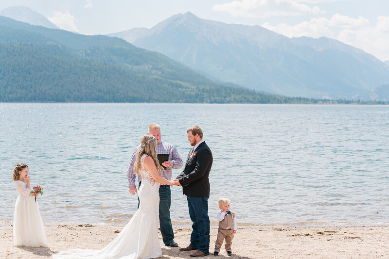 A couple stands with their two small children and officiant next to a lake and mountains