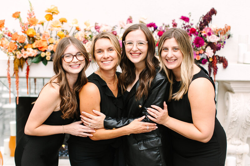 Four SoHo63 team members pose beneath a floral-covered archway at the entrance of SoHo63, a Chandler wedding and event venue. All are dressed in black and smiling, standing in front of the venue’s signature arched doors framed by lush white blooms and greenery.