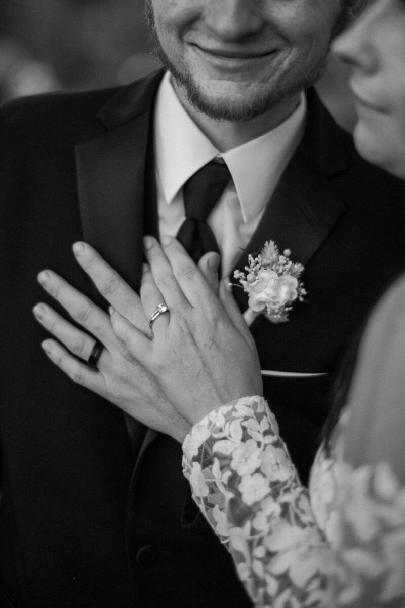A close-up of two hands, one with a ring, gently touching a man's chest, showcasing elegance and tenderness in a black and white setting.