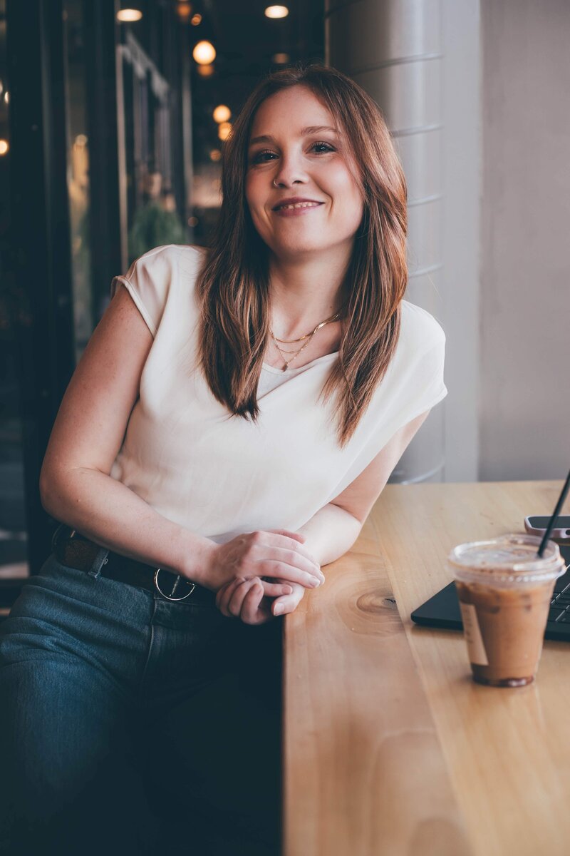 A woman with long hair, wearing a black blazer and ripped jeans, sits on an antique chair with one foot on a small wooden box, in a room with wooden flooring of an agency offering branding and web design for female entrepreneurs.