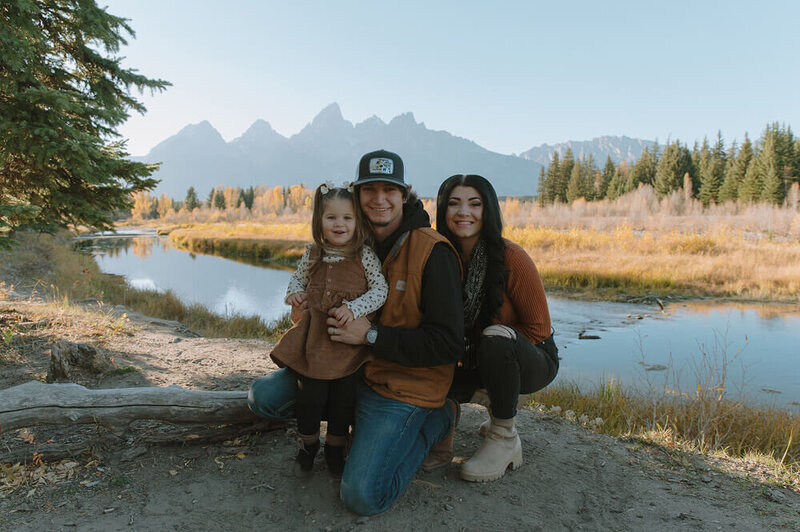 Kneeling close to the ground, a wife holds her husband from behind as he holds their daughter from behind. The outline of the Grand Tetons are behind them.