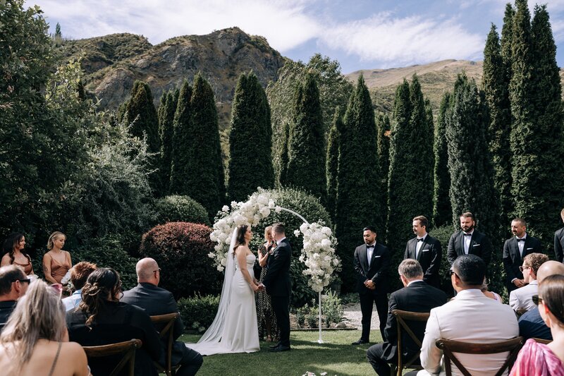 a wide photo of the winehouse queenstowns ceremony lawn on a summer day with white floral arch