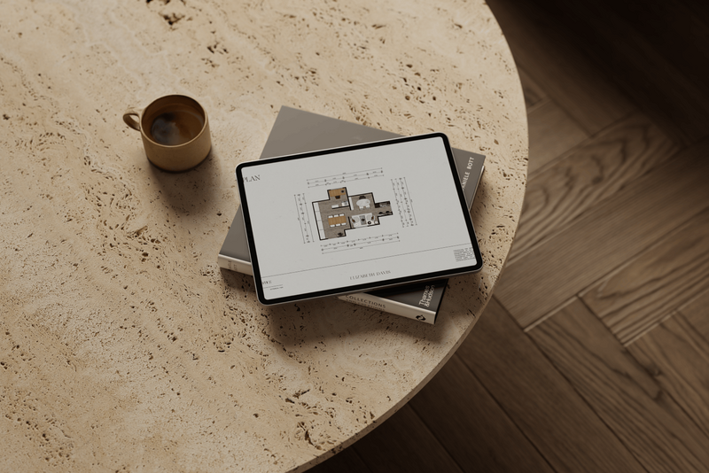 iPad placed atop a book on a travertine coffee table, accompanied by a coffee cup, displaying a 2D floor plan of an interior design studio.
