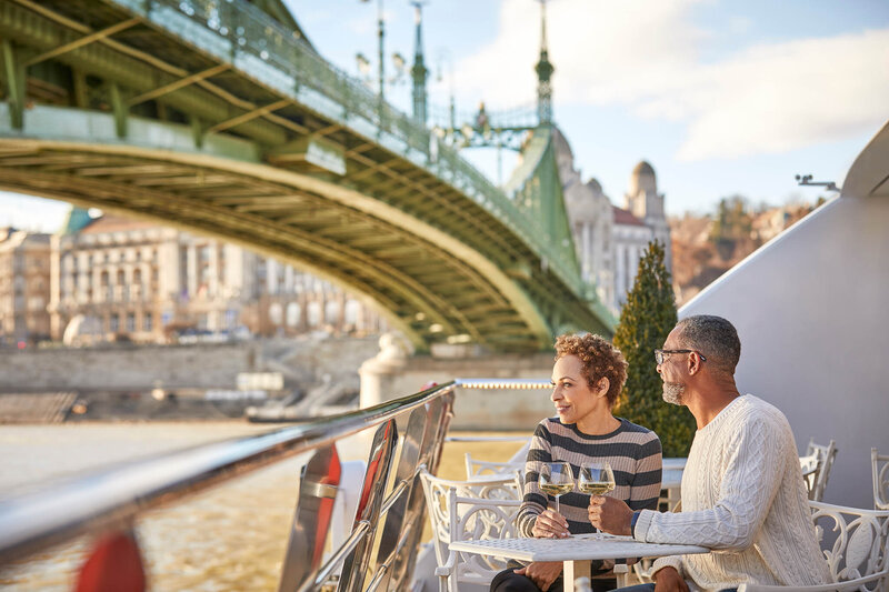 A couple sits at a small outdoor table on a boat deck, enjoying drinks while overlooking a river and a large green metal bridge. The sun is warm and low, illuminating the historic buildings along the riverbank in the background.
