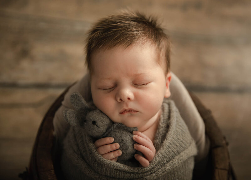 Newborn boy holding a felt gray rabbit as he sleeps inside a wooden bucket.