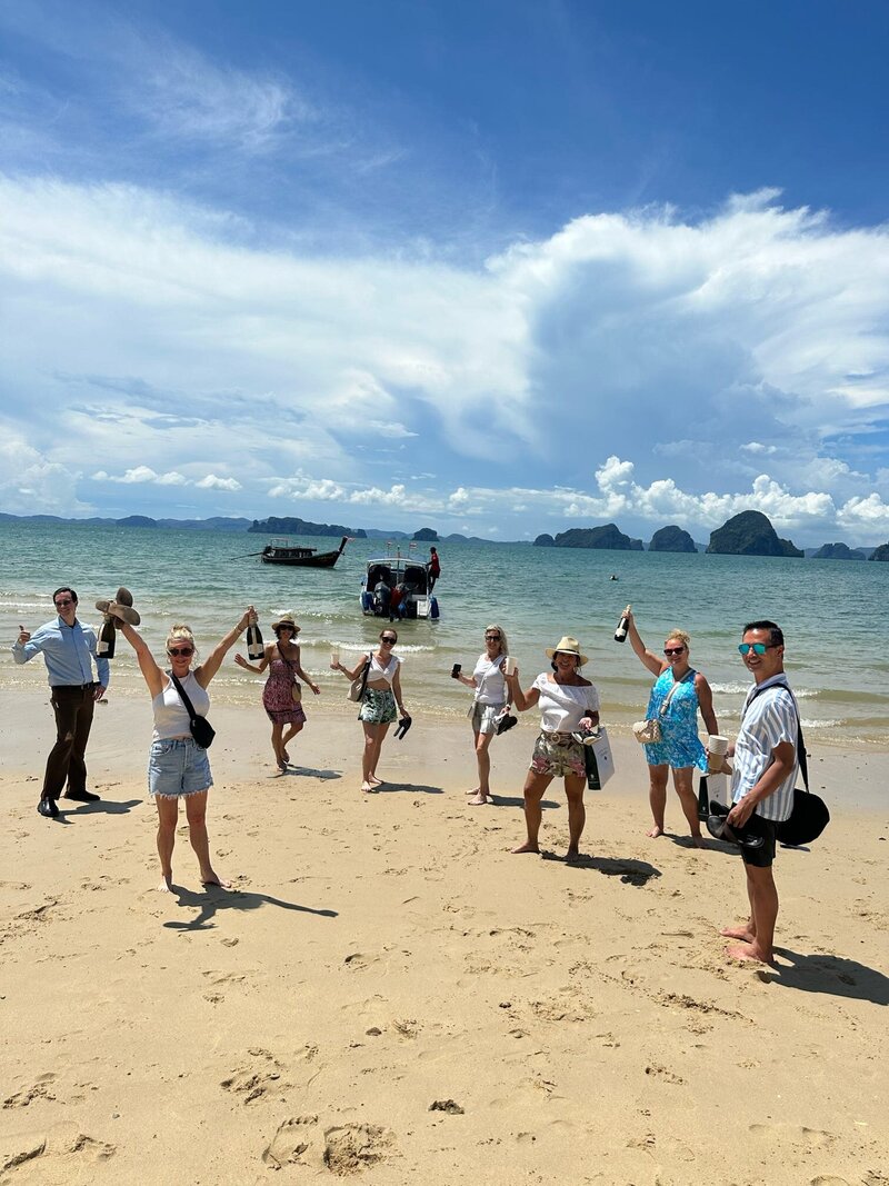A group of men and women standing on the beach, each holding a bottle and raising it in celebration. In the background, a boat is anchored near the shore, with distant islands and a partly cloudy sky on the horizon.