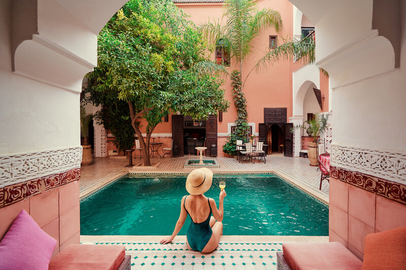 A person in a swimsuit and sun hat sits by a beautiful, tiled pool, holding an ice cream. The setting is a courtyard with arches, lush greenery, and peach-colored walls, creating a serene, luxurious atmosphere.