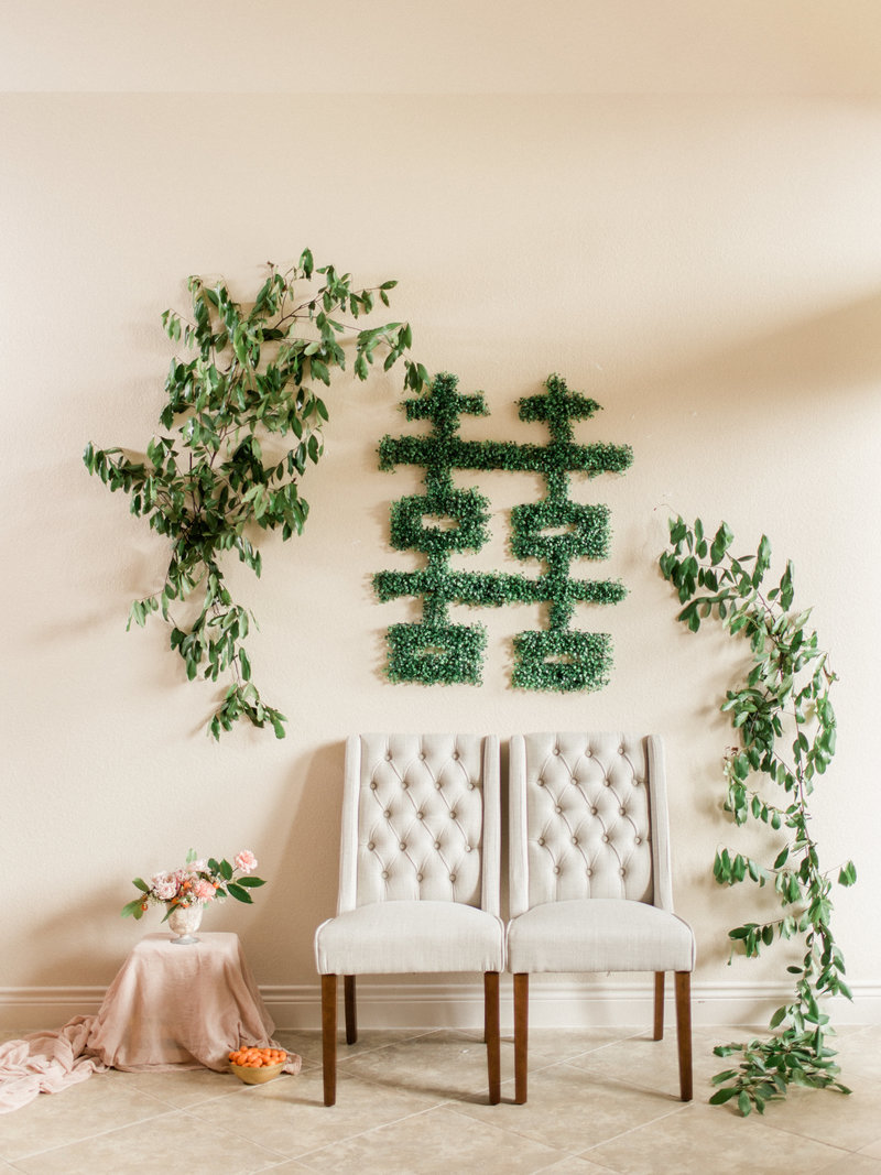 Two chairs sit in front of a wall with the Chinese Double Happiness symbol in greenery for couple's tea ceremony