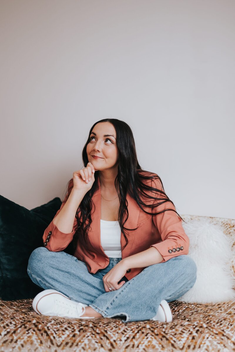Woman with long, dark hair smirks while looking off-camera wearing a salmon-colored blazer and jeans.