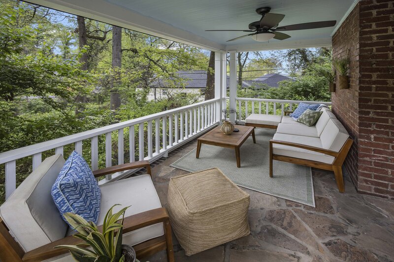 A photograph of a room Alicia designed: a blue-ceilinged porch with a sectional, coffee table, arm chair, and footrest poof. You can see houses and trees in the distance.