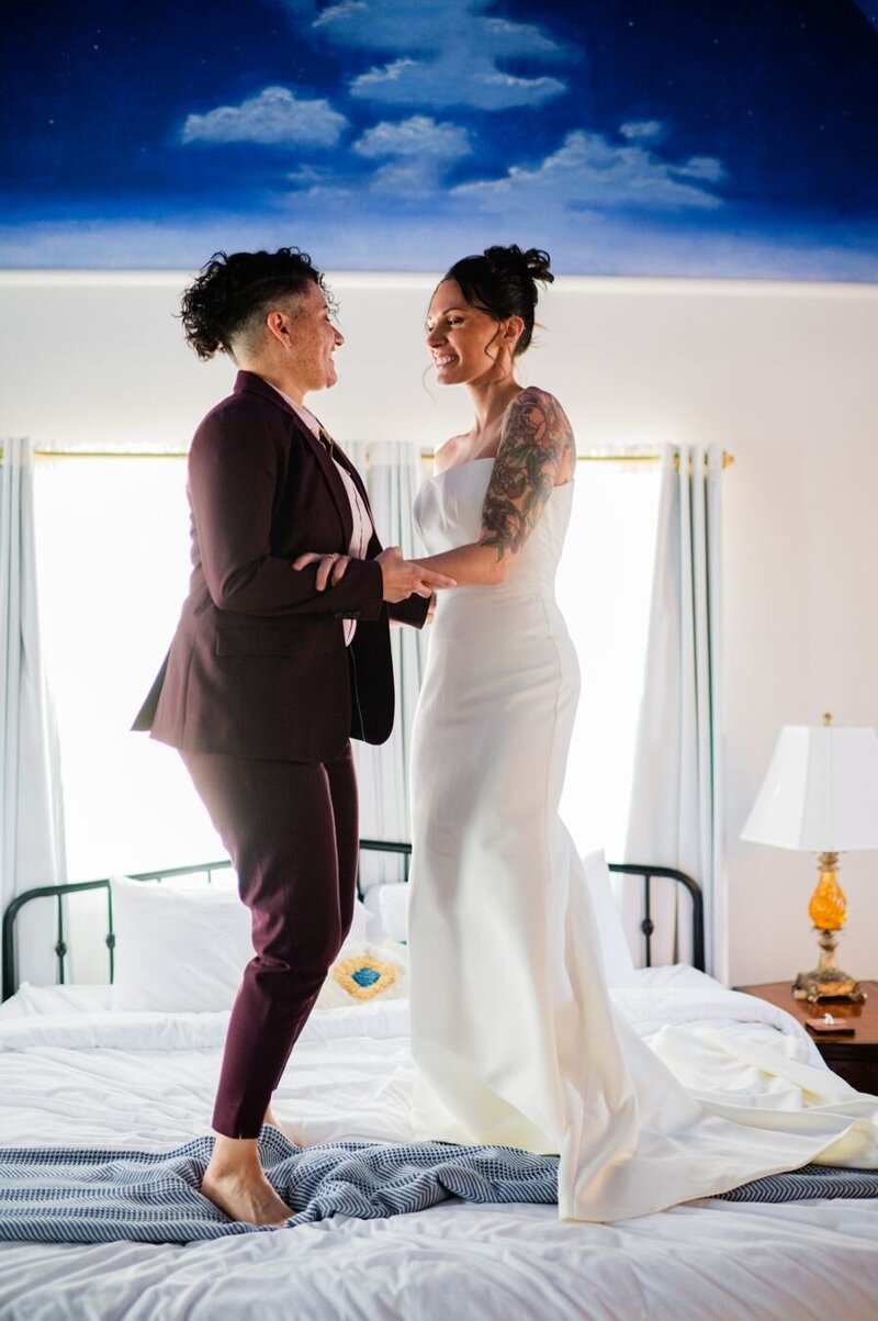 A couple jumps on a bed, barefoot, holding both hands. One wears a white wedding gown, with hair in an updo, and a visible tattoo sleeve. The other wears a burgundy two-piece suit.