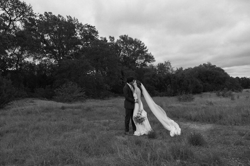 Bride and groom kissing in a field while the brides veil blows off behind her. 