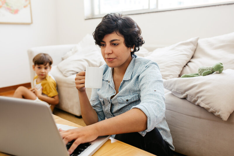 Non-profit remote worker and mom, parent works from home on a laptop and drinks coffee as her son plays nearby 