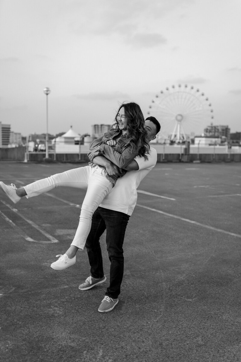 J picking up J from behind and swinging her around, laughing, on the rooftop of a parking garage with the ferris wheel in the background
