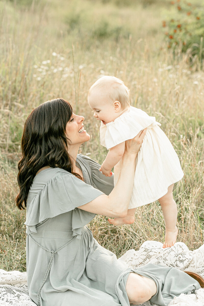 Mom in sage green dress holding up one year old baby girl in ivory dress. They are both smiling really big at each other. Sitting in a field of tall grasses. Portland Family Photography session outdoors.
