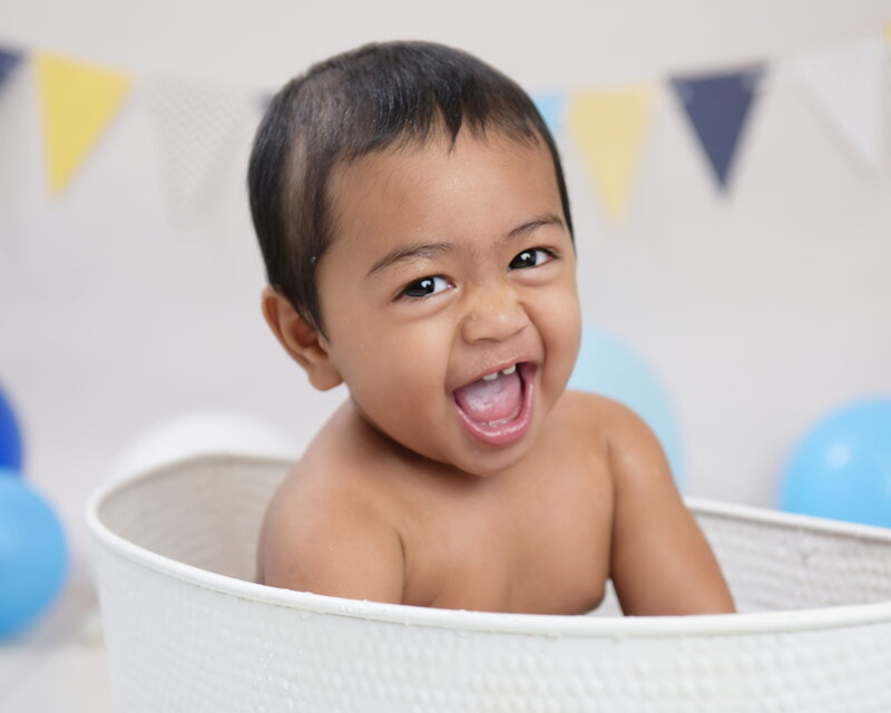 A baby boy laughing joyfully while sitting in a white tub, surrounded by blue and yellow balloons with bunting in the background.