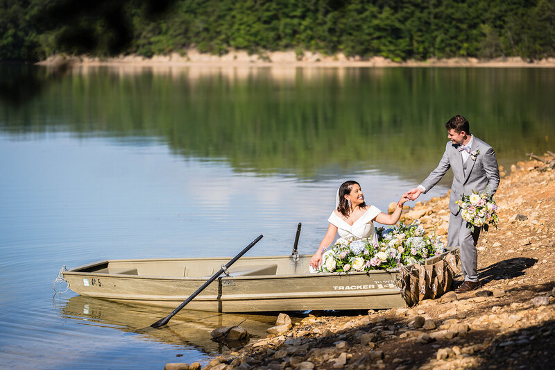 A groom helps a bride into a rowboat adorned with pastel flowers while holding his bride's bouquet as the pair smile at one another at Carvin's Cove in Roanoke, Virginia on their elopement day.