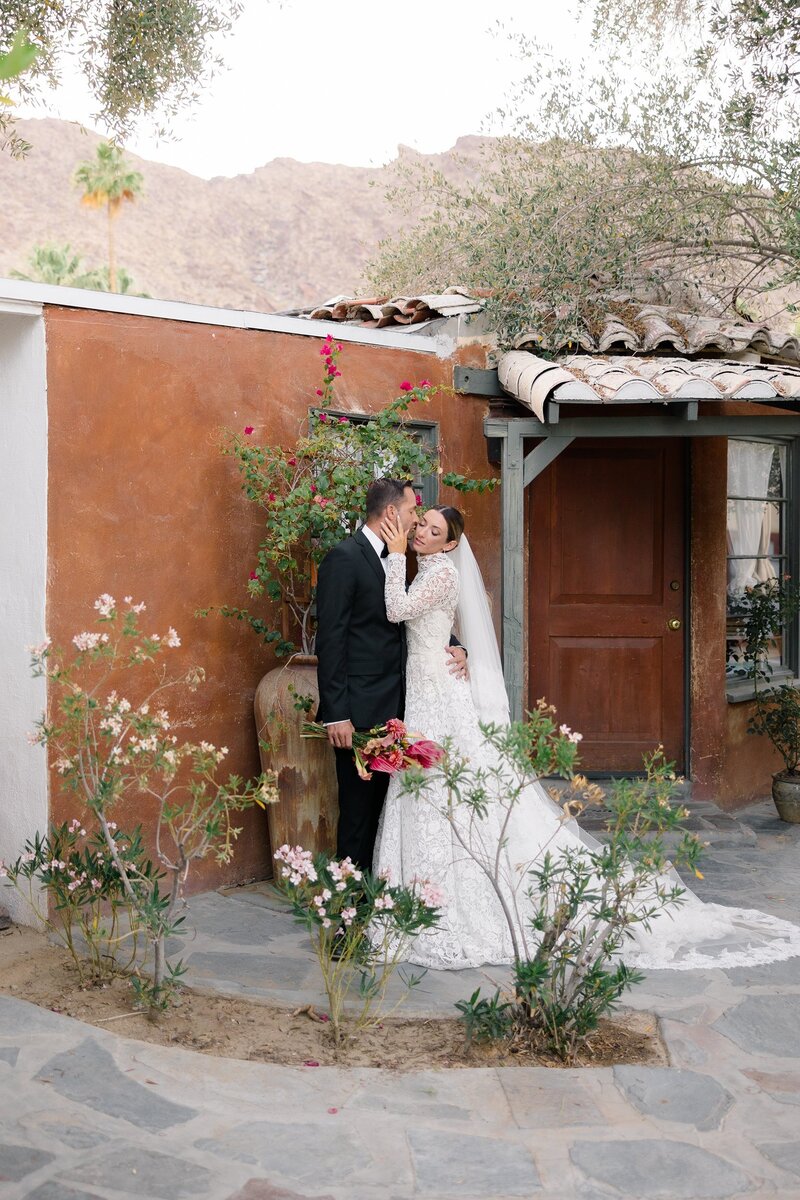 Bride and groom posing in front of a rustic home with mountain backdrop.
