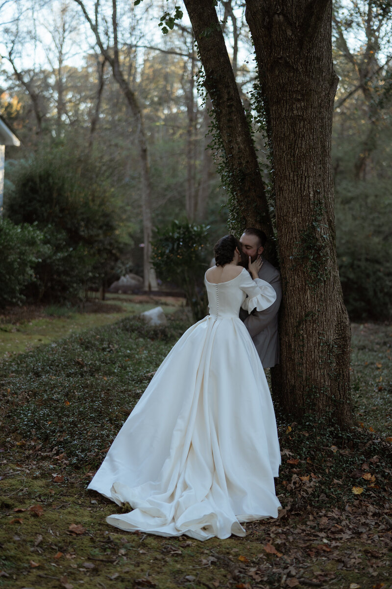 Bride in an elegant, long-sleeved satin gown with a dramatic train, sharing an intimate kiss with the groom against a tree in a serene, wooded outdoor setting.