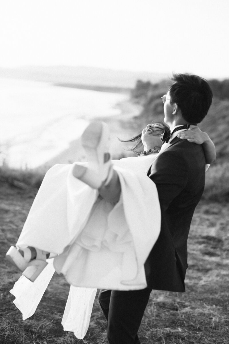 The groom holds the bride in his arms as she kicks up her leg and laughs near the cliffs of Dos Pueblos Orchid Farm in Santa Barbara, in black and white shot on film by Southern California film photographer Megan Lynn of My Sun and Stars Co.
