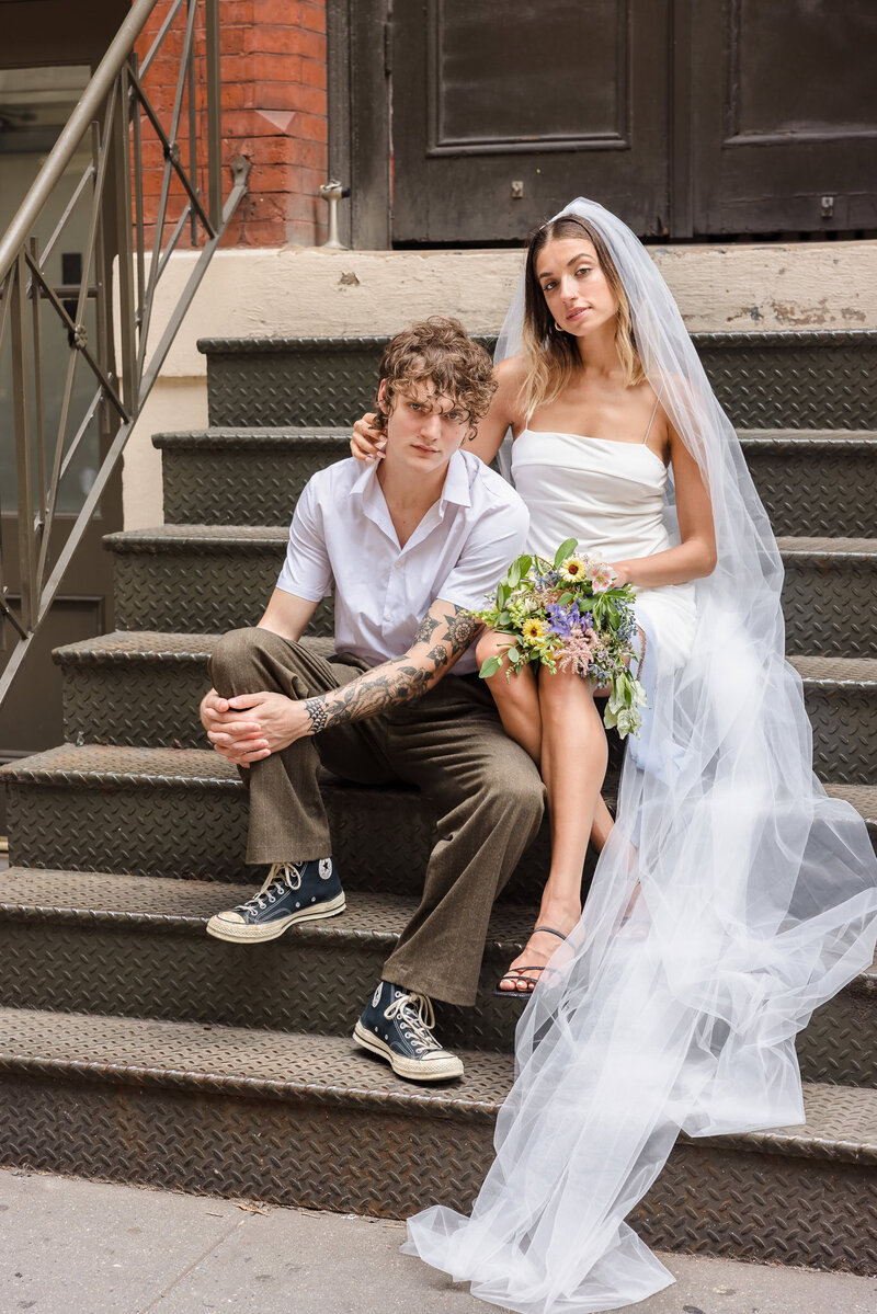 Modern bride and groom pose on SoHo brownstone steps in NYC, her veil cascading down the stairs as she holds a vibrant bouquet, photographed by NYC wedding photographer Heritage House.