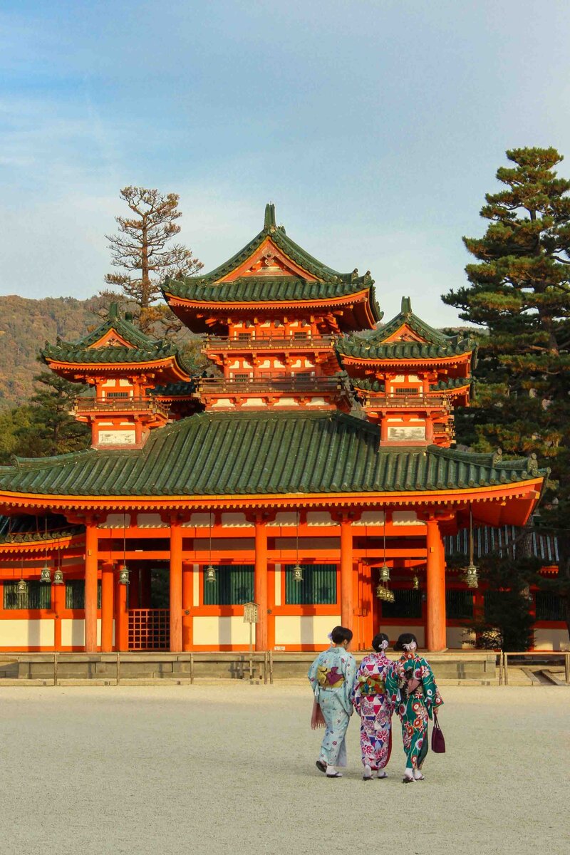 Three women in colorful kimono walking towards the bright vermilion and green-roofed Heian Shrine, surrounded by trees in Kyoto, Japan ©Stephanie Dosch | theViatrix Luxury Travel