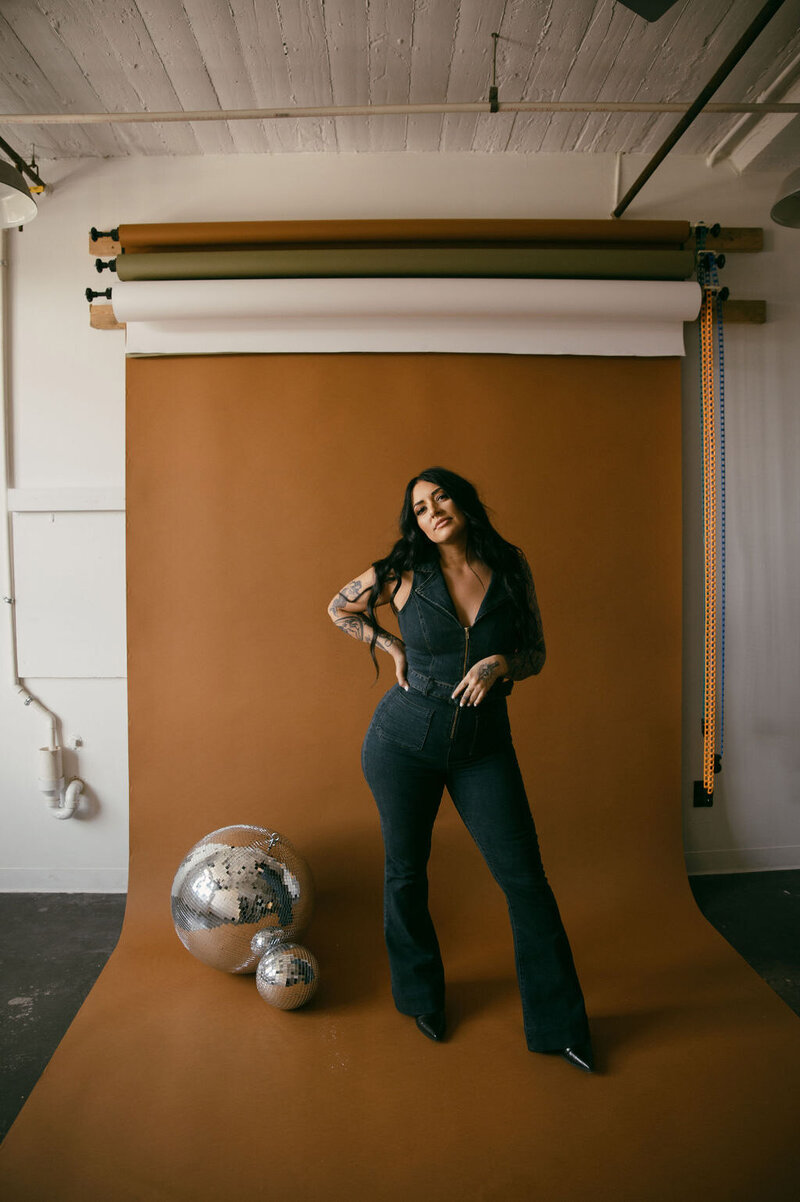 Morgan Rindahl standing on an orange photo backdrop next to a large disco ball wearing a denim jumpsuit