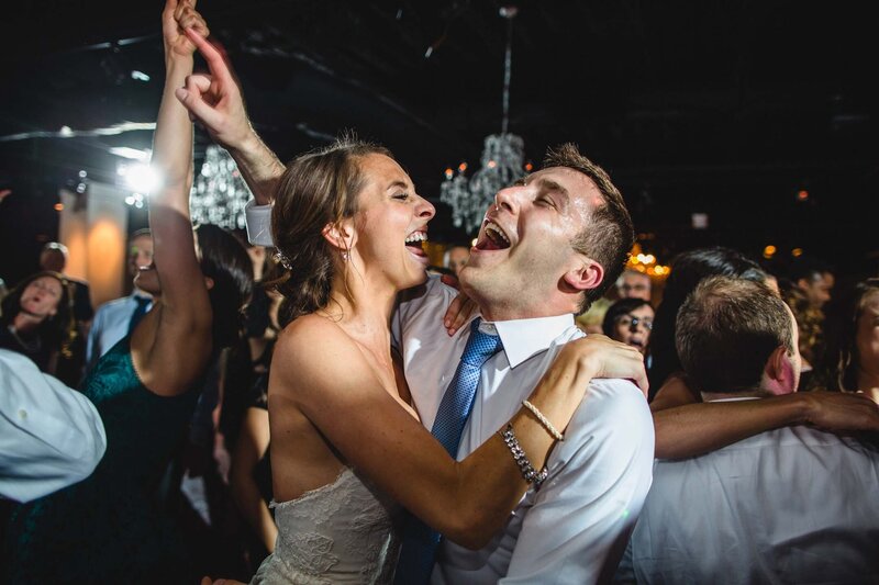 A newlywed couple shares a kiss after a Jewish wedding at the Boathouse at Confluence Park surrounded by guests holding sparklers at night, creating a magical and festive atmosphere. The bride's curly hair and the groom's beard are softly illuminated by the warm light of the sparklers.