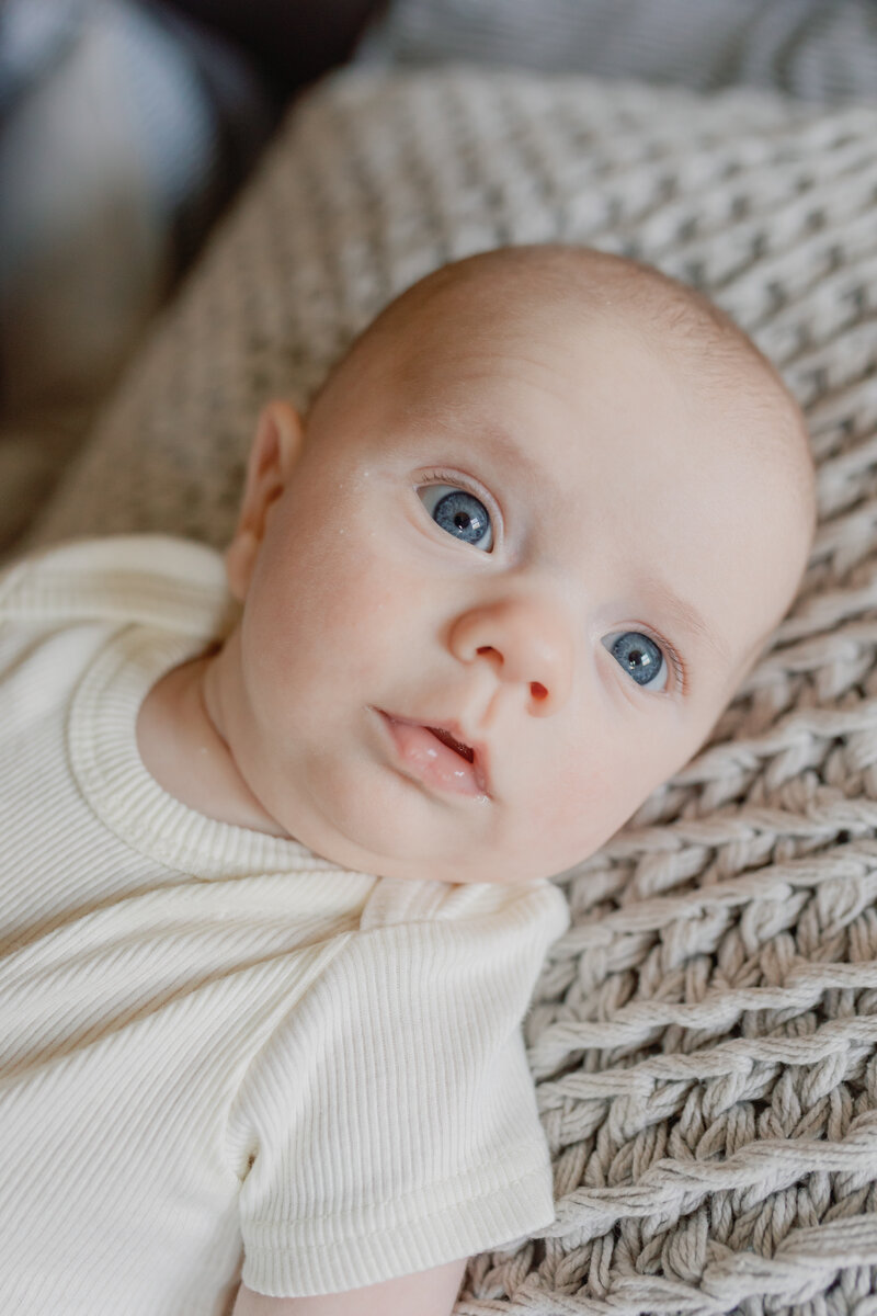 A close-up newborn portrait featuring a baby with bright blue eyes gazing curiously at the camera. The baby, dressed in a soft cream-colored onesie, lies on a textured knit blanket, bathed in natural light. This timeless image captures the innocence and wonder of early childhood, a signature of KCP Families' storytelling photography.