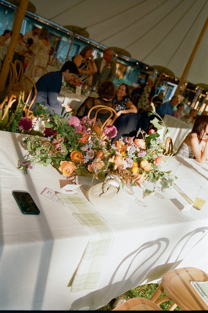 Reception guest table with a garden-inspired centerpiece of orange ranunculus, pink roses, and yellow blooms with greenery, designed by Summer Robbins Flowers in Central Oregon