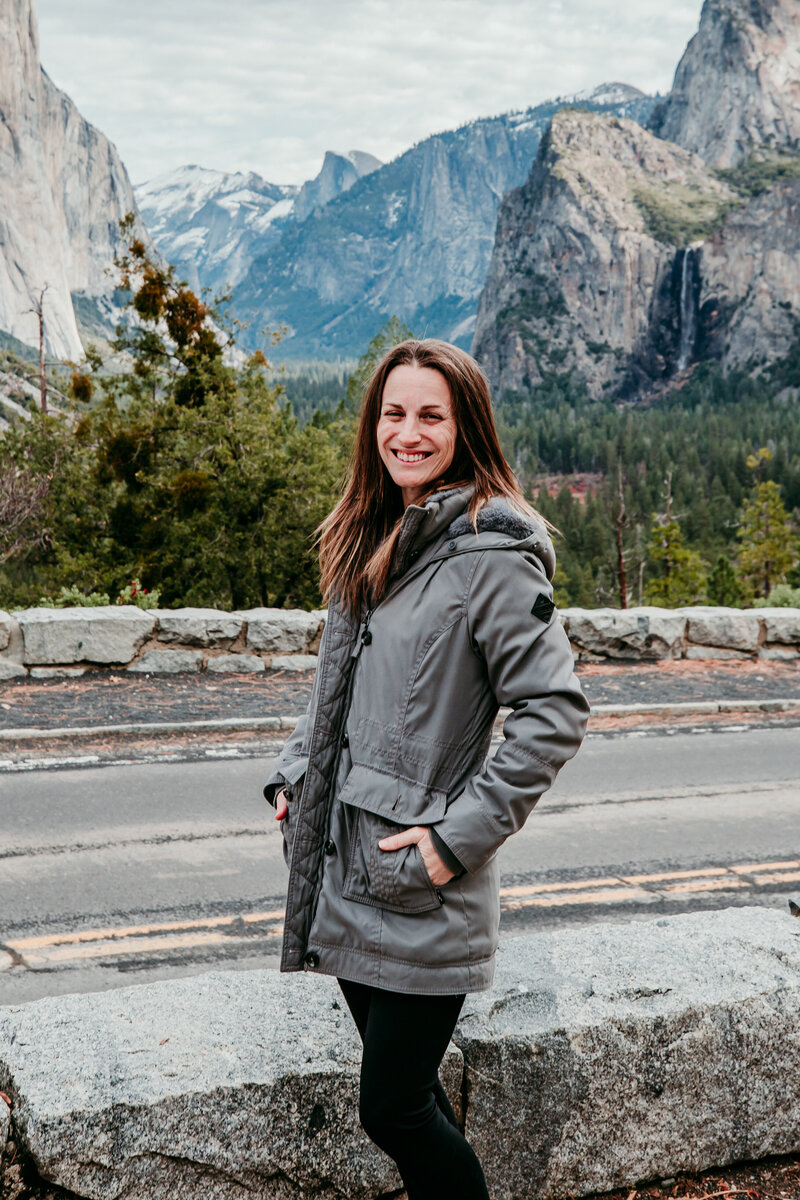 Kristin Smith, Lake Tahoe and Yosemite elopement photographer, smiling at Tunnel View in Yosemite National Park with granite cliffs and pine trees in the background.