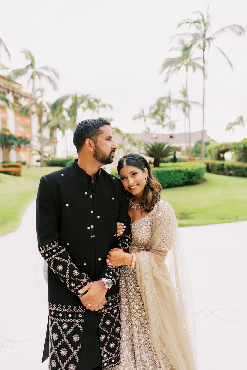 A couple dressed in traditional Indian wedding attire pose outdoors, with the bride resting her head on the groom’s shoulder. The groom wears a black sherwani, and the bride wears a light-colored embellished saree.