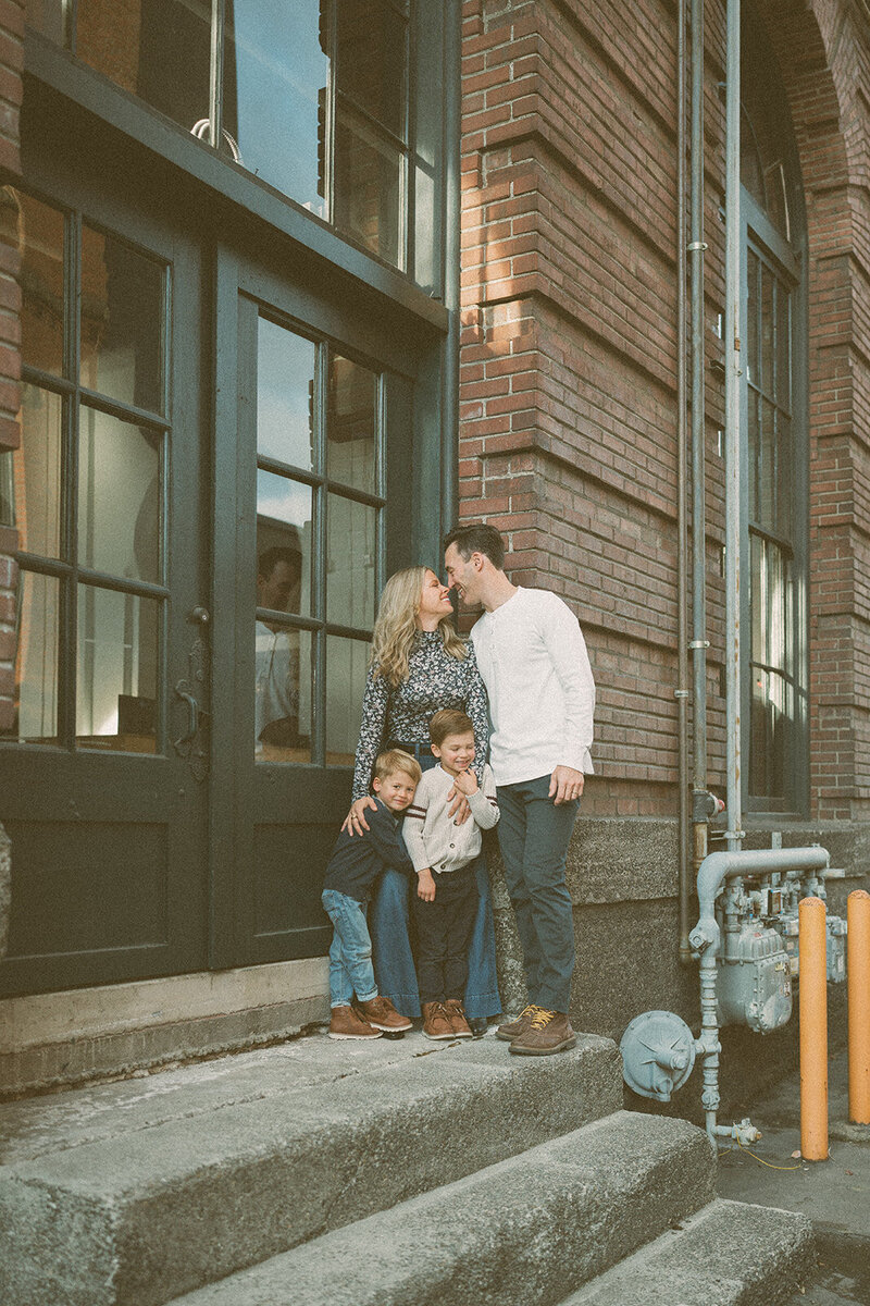 A candid family moment on the steps of a brick building during the first location of their two-part documentary session. The parents lean in together while their two boys gather close, creating an easy, connected scene.