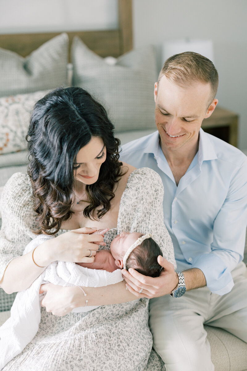New parents snuggled on a bed with their newborn baby during a relaxed, natural-light Charlotte photo session.