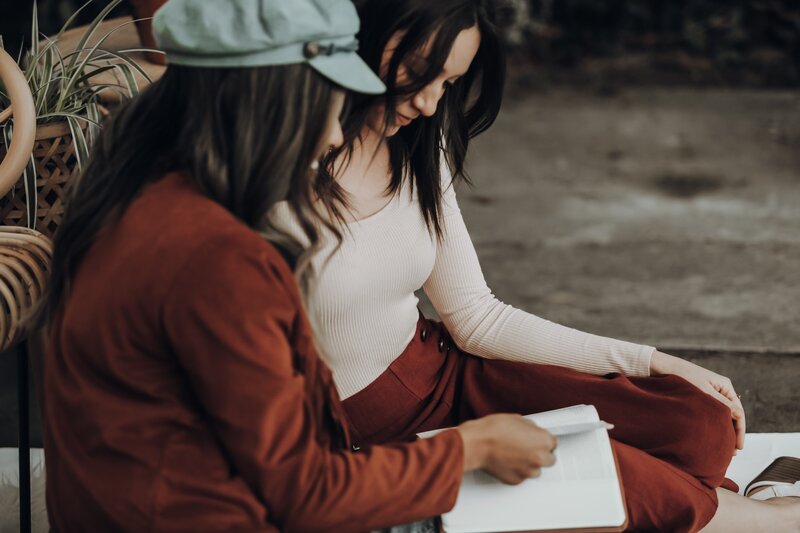 Two female friends wearing a hat and dark clothes both looking down at a book on one of the women's lap