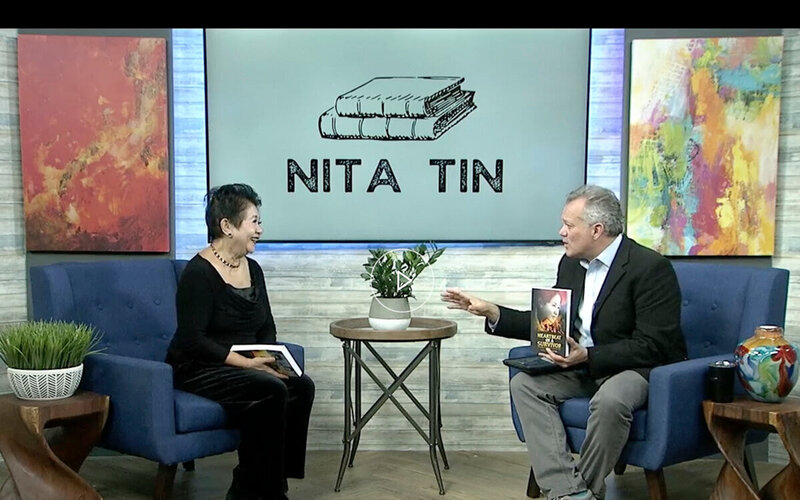Nita Tin interviewed on Channel 9 Chattanooga’s This and That series. She is seated in a studio blue chair next to host James Howard, who holds her book Heartbeat of a Survivor. A small table with a plant sits between them, with a backdrop screen displaying her name and book graphic.